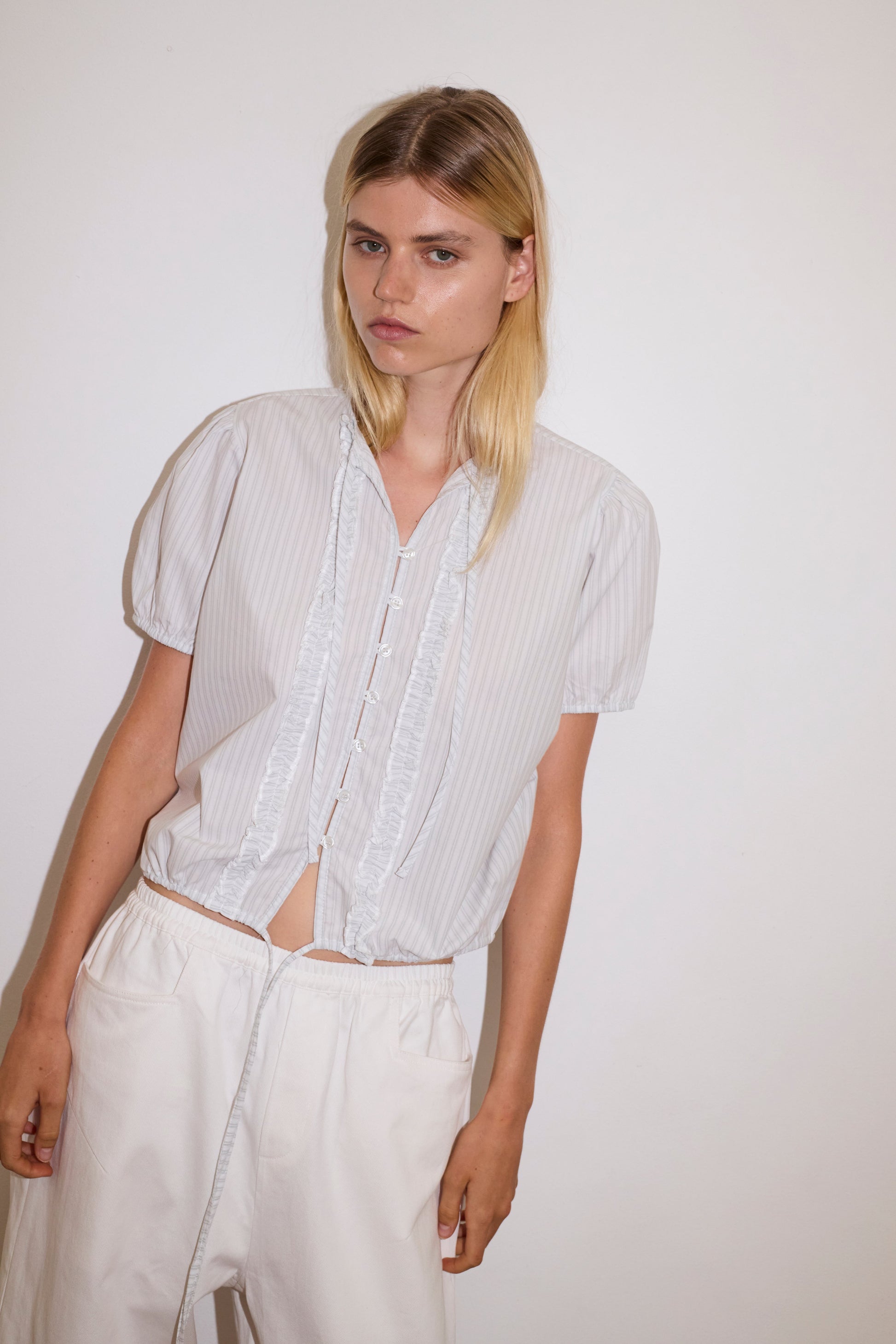 A young person with straight blonde hair wears the Double Frill Top - Sage Stripe by Deiji Studios, featuring subtle stripes and ruffle details, paired with loose white drawstring pants against a plain white background.
