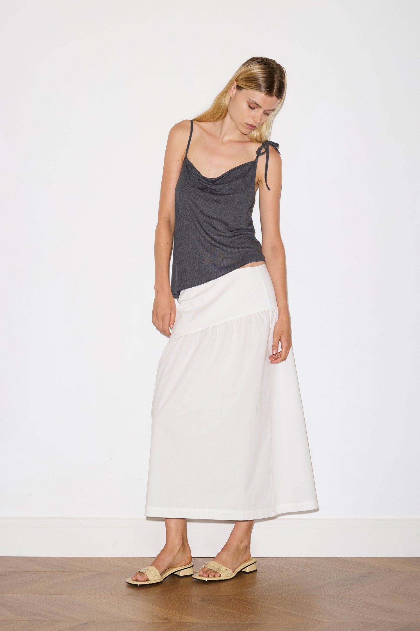 A woman stands indoors against a plain white wall and wooden floor, wearing the Angle Skirt—a long white skirt—paired with a dark gray sleeveless top with thin straps and beige sandals. She looks down, her head gently tilted.