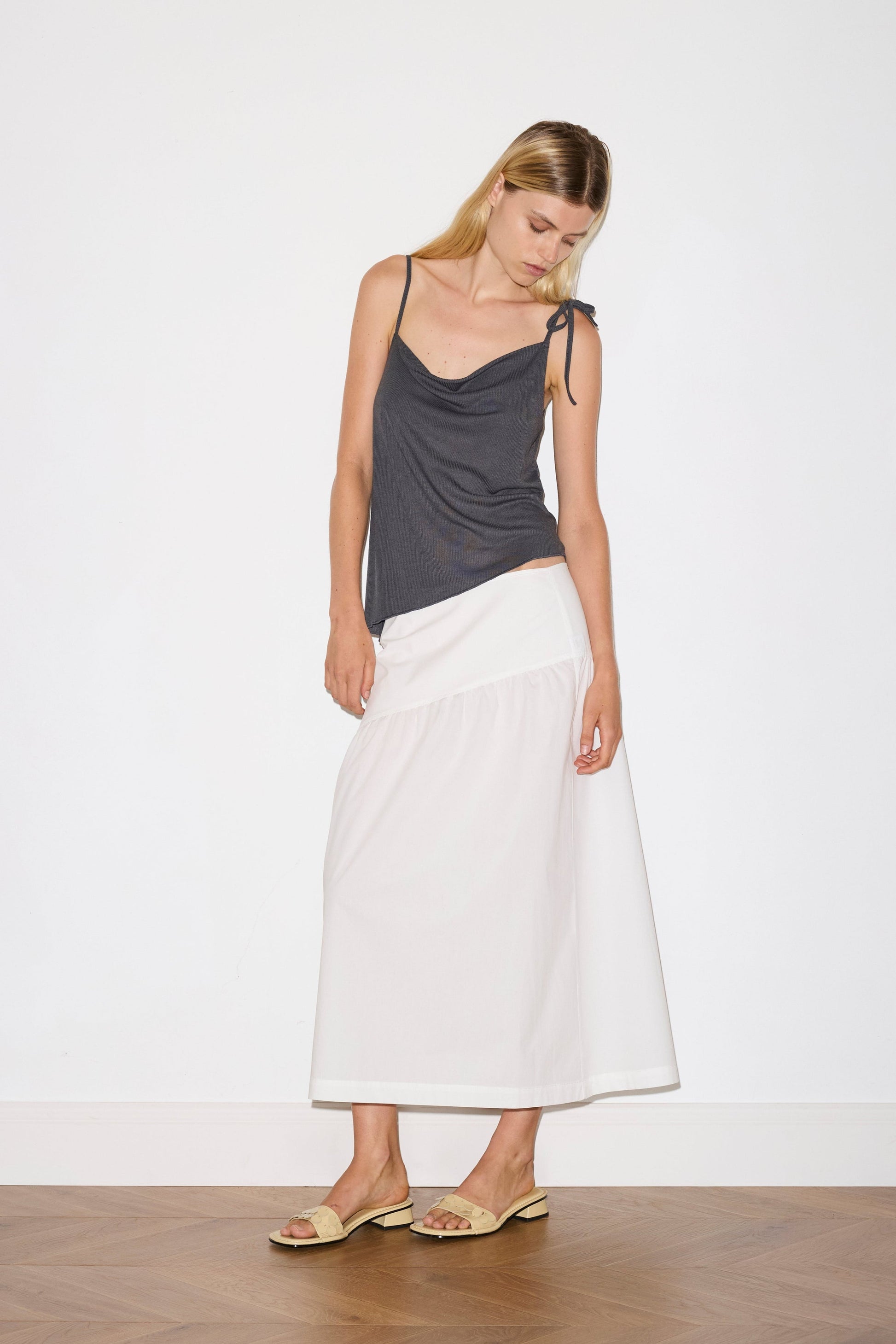 A woman stands indoors against a plain white wall and wooden floor, wearing the Angle Skirt—a long white skirt—paired with a dark gray sleeveless top with thin straps and beige sandals. She looks down, her head gently tilted.