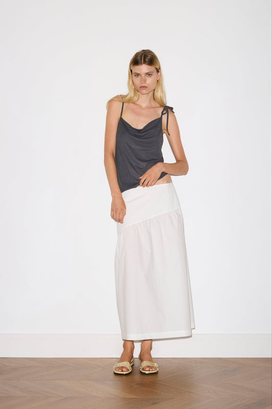 blonde female model wears a sheer fine knit relaxed tank with a fold over cowl neckline and low draping back, features self fabric rouleau straps, baby lock hem finishes and a asymmetric bottom detail, worn with a midi length white skirt 