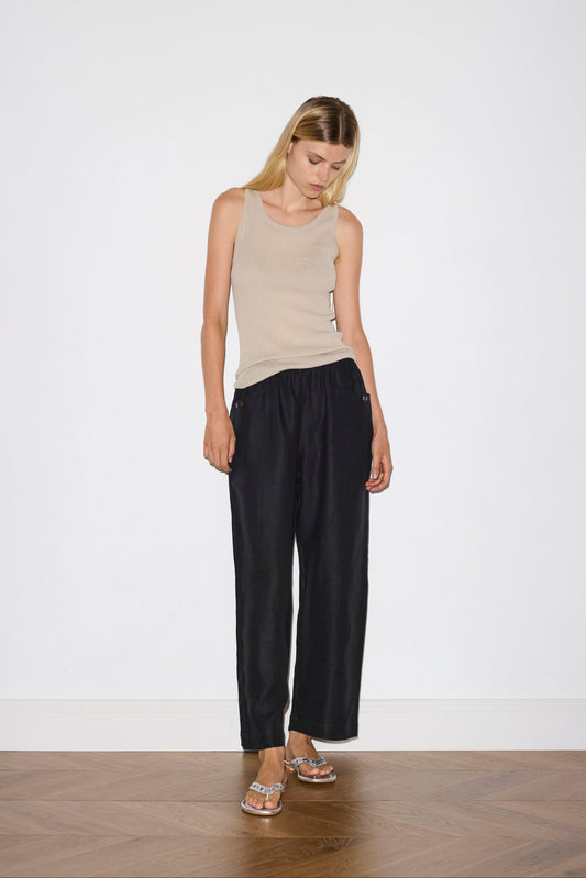 A woman with long blonde hair stands indoors against a plain white wall, wearing black Linen Square Pocket Pants, a beige sleeveless top, and sandals. She looks down with her arms relaxed at her sides.