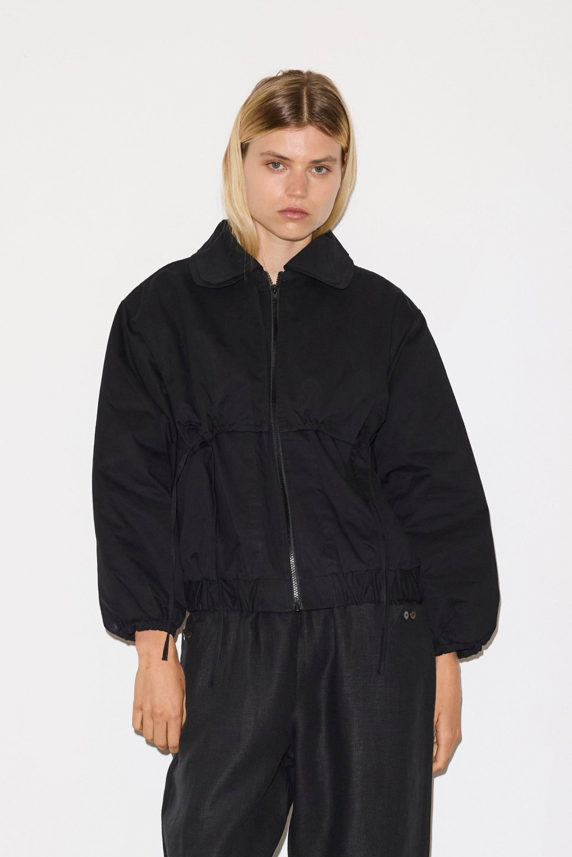 A woman with straight blonde hair models the 0 Double Layered Jacket in black with a collar and loose black pants, standing against a plain white background.