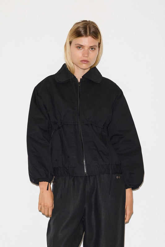 A woman with straight blonde hair models the 0 Double Layered Jacket in black with a collar and loose black pants, standing against a plain white background.