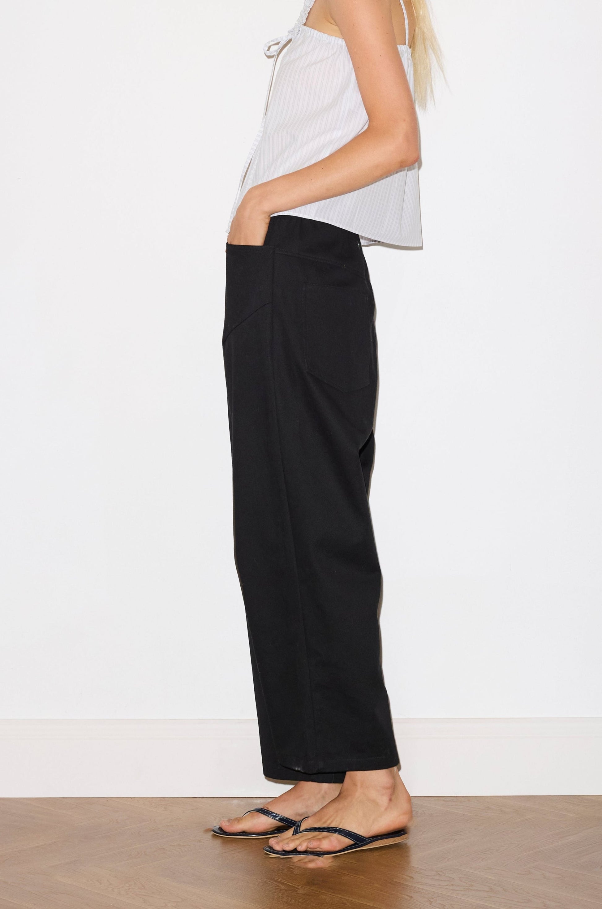 blonde haired female model wears a black Heavy- weighted relaxed-fit pant featuring a wide-leg silhouette, elasticated waistband, and square pocket detail, worn with a sage and white striped a relaxed-fit top featuring self-fabric ruffles along the straps and center front, with a tie front detail. designed with elasticated gathering for ease and comfort