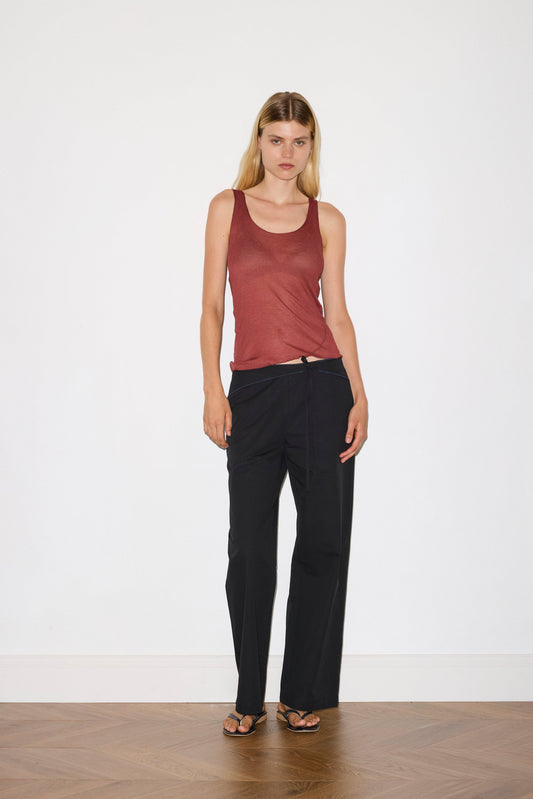 A woman with long blonde hair and a neutral expression stands indoors against a white wall, wearing the Belted Pant - Black with a red sleeveless top and sandals. The wooden floor beneath her features a herringbone pattern.