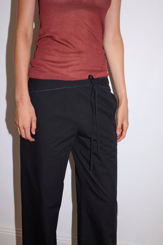 A person wearing a sleeveless, ribbed, rust top and loose-fitting black Belted Pant stands against a plain white wall with their left hand in their pocket.