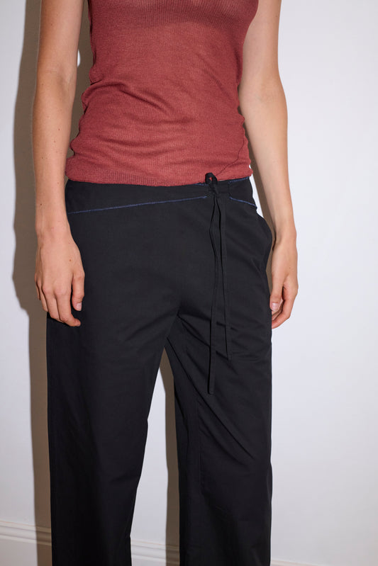 A person models the 0 Belted Pant - Black, a relaxed fit style with a drawstring, paired with a sleeveless, ribbed reddish-brown top. Photo shows torso and upper legs against a plain white wall.