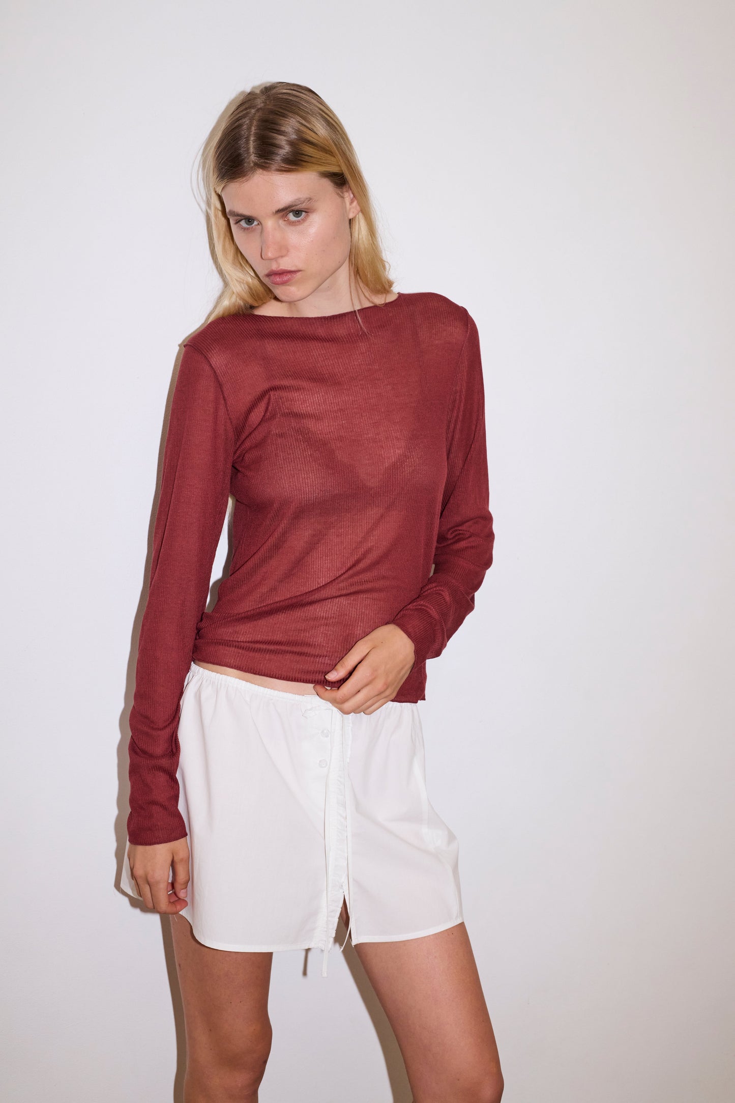 blonde female models wears a red a sheer fine knit long sleeved top with exposed seams and tag, featuring baby locked finishes and a relaxed fit, worn with a white simple mini skirt designed to sit at low waist features adjustable tie and ruffle edge on side with button detail