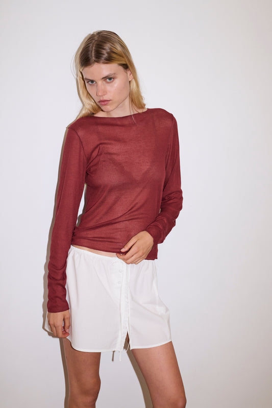 blonde female models wears a red a sheer fine knit long sleeved top with exposed seams and tag, featuring baby locked finishes and a relaxed fit, worn with a white simple mini skirt designed to sit at low waist features adjustable tie and ruffle edge on side with button detail