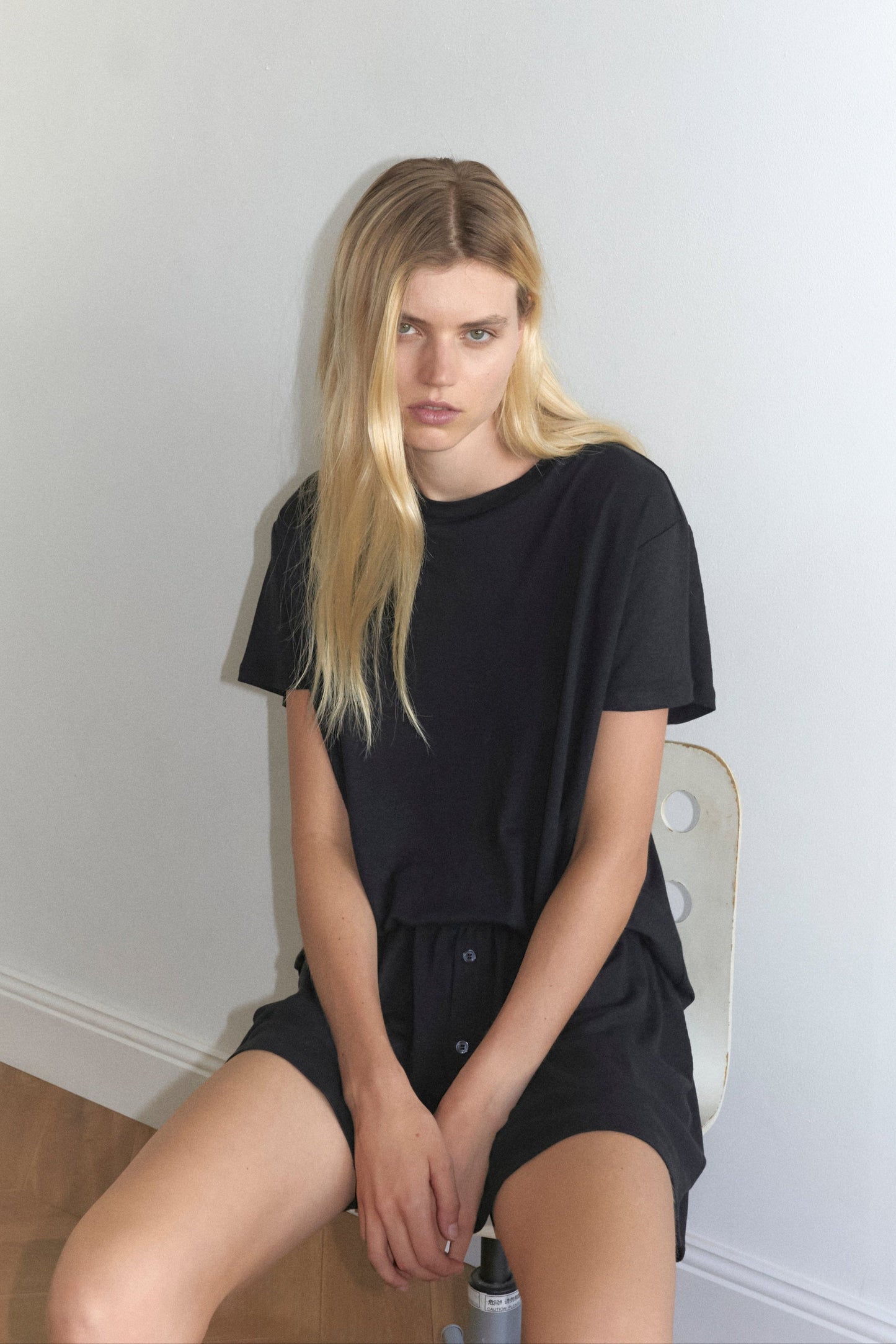 A woman with long blonde hair sits on a chair against a white background, wearing Deiji Studios Soft Short in black paired with a matching black t-shirt, her hands are rested crossed on her lap.