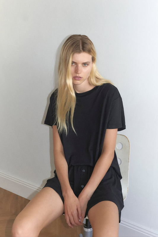 A young woman with long blonde hair sits on a white chair against a light-colored wall, wearing Deiji Studios’ soft t shirt - black and matching shorts, looking slightly down with a neutral expression.