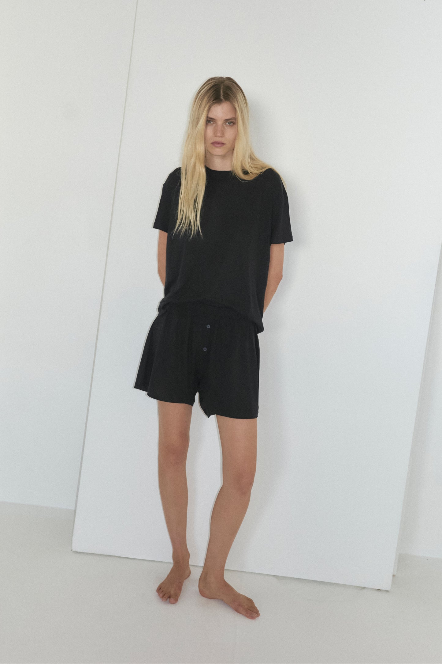 A young woman with long blonde hair stands against a plain wall, wearing the Deiji Studios Soft T Shirt in black and matching shorts, gazing upwards towards the camera with a neutral expression.