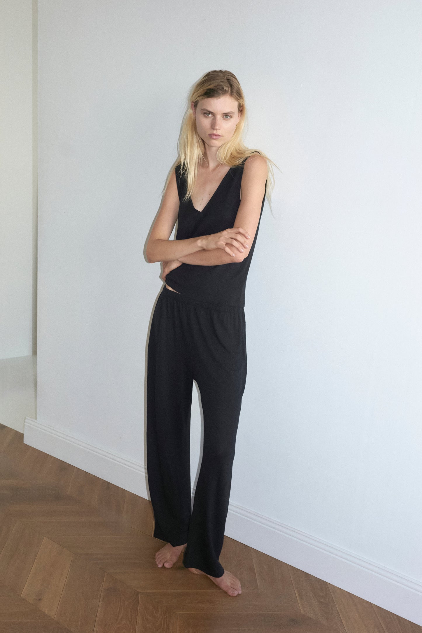 A woman with long blonde hair stands barefoot on a wooden floor, leaning against a white wall in Deiji Studios’ soft pant - black and a sleeveless black top, gazing at the camera with a neutral expression.