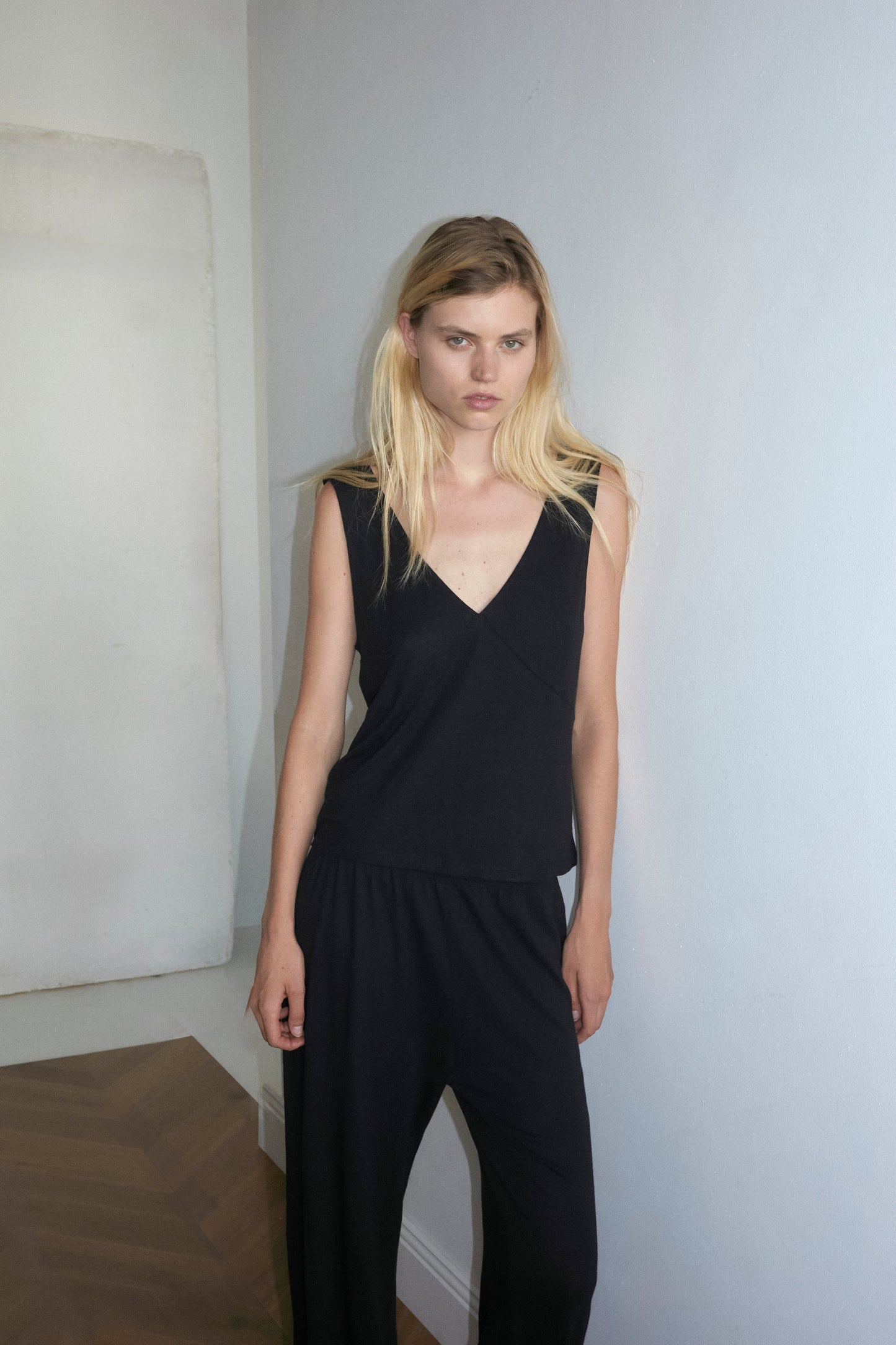 A blonde-haired person wears Deiji Studios soft tank - black, paired with a black pant and standing and leaning against a white wall and looking at the camera with a neutral expression.