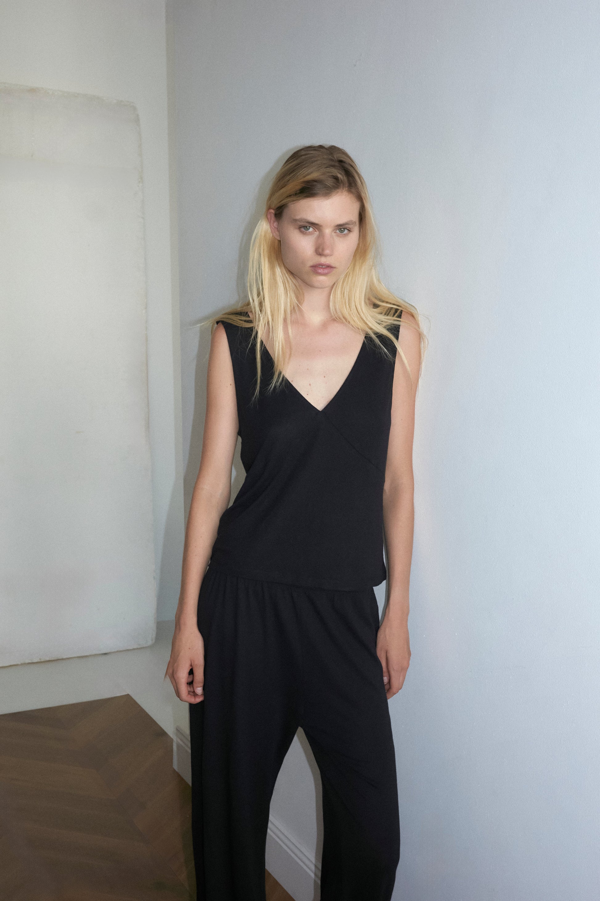 A woman with long blonde hair stands barefoot on a wooden floor against a white wall, wearing Deiji Studios Soft Pant and a sleeveless black top. Her arms are rested beside her and has a neutral expression.
