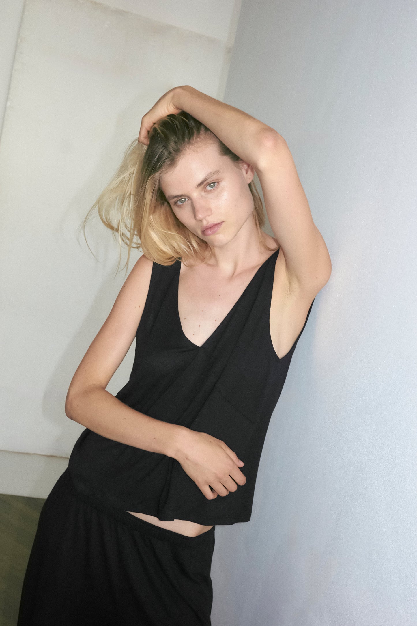 A blonde-haired person wears Deiji Studios soft tank - black, standing indoors against a light blue wall with one arm raised, touching their head, and looking at the camera with a neutral expression.