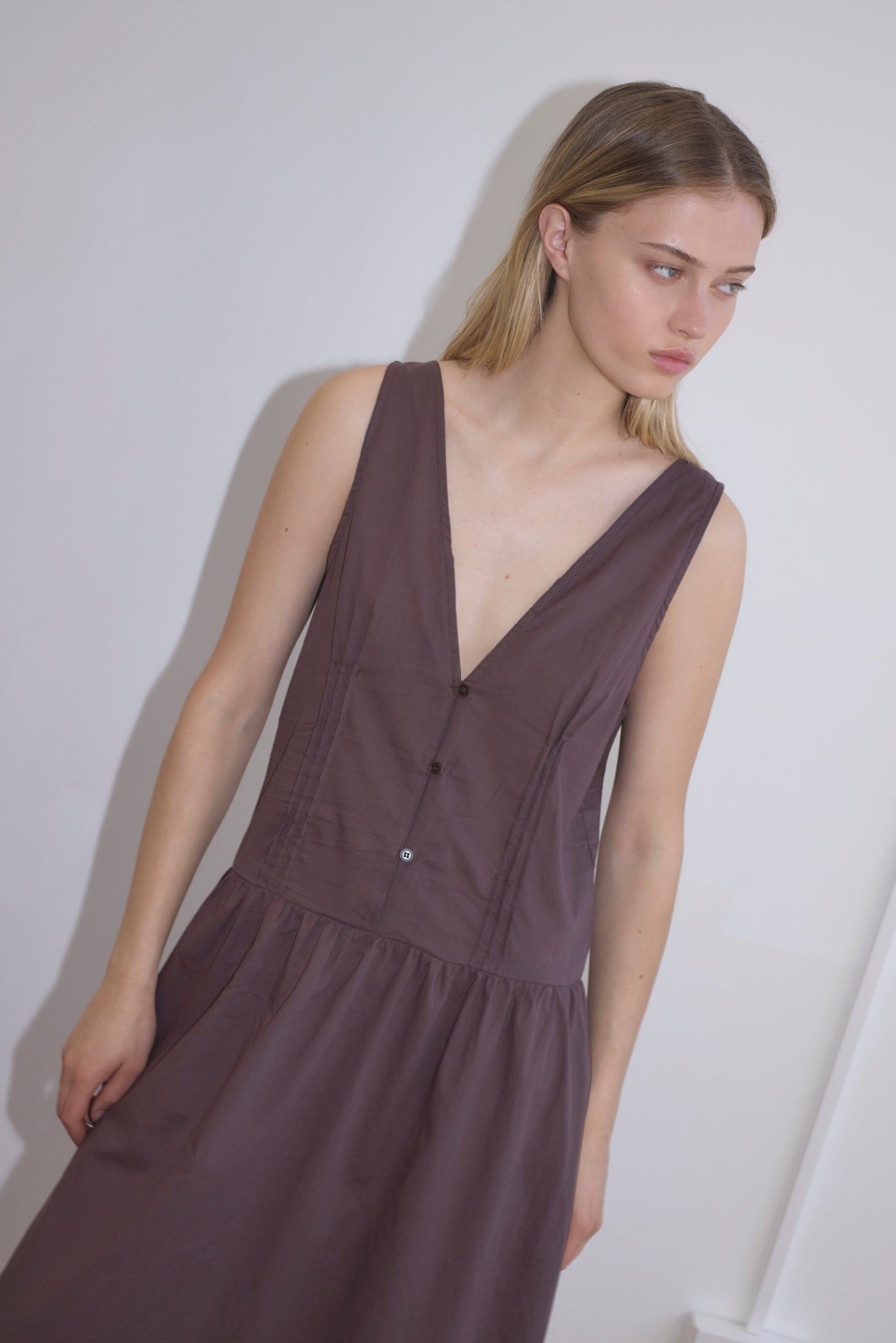 A young woman with light brown hair wears the Pintuck Dress - Raisin, a sleeveless, deep V-neck, mid-length dark purple dress, standing against a plain white wall and gazing to the side with a neutral expression.