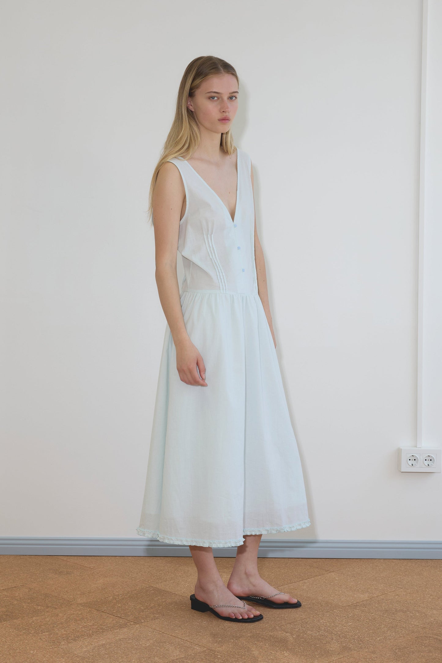 A blonde model wears the Pintuck Dress, a sleeveless white midi with V-neckline, button front, and flowy lace-trim skirt, paired with black chain-detail flat sandals.