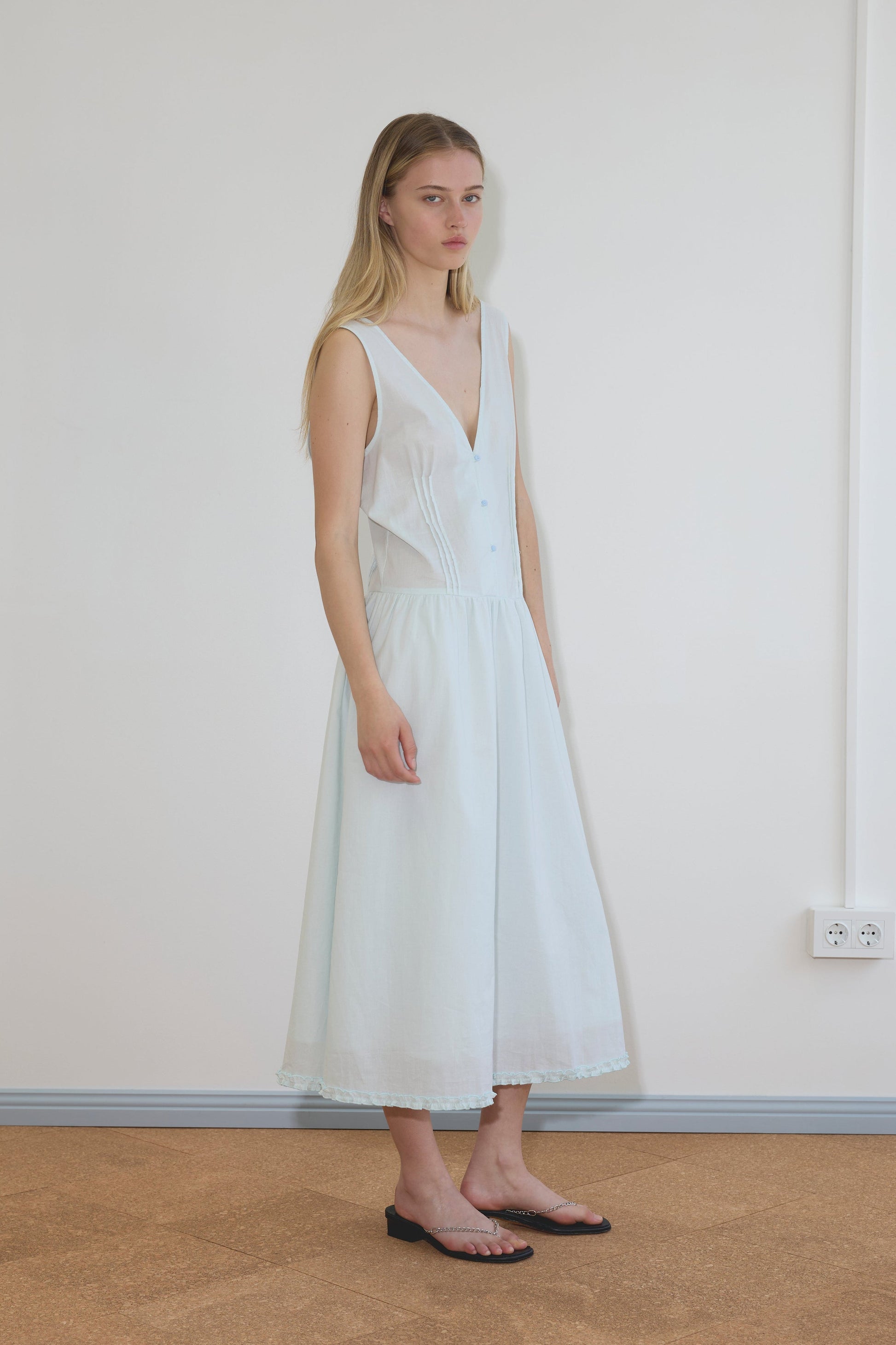 A blonde model wears the Pintuck Dress, a sleeveless white midi with V-neckline, button front, and flowy lace-trim skirt, paired with black chain-detail flat sandals.