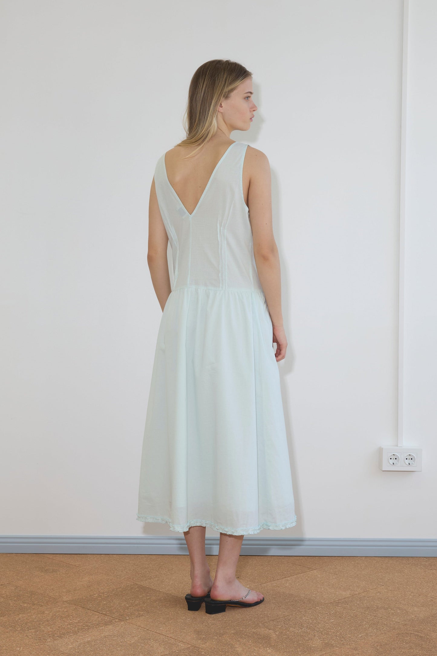 A woman with shoulder-length blonde hair and black sandals stands indoors, facing away, wearing the Pintuck Dress - Mint by Deiji Studios. The room has white walls, a carpeted floor, and a visible power outlet.