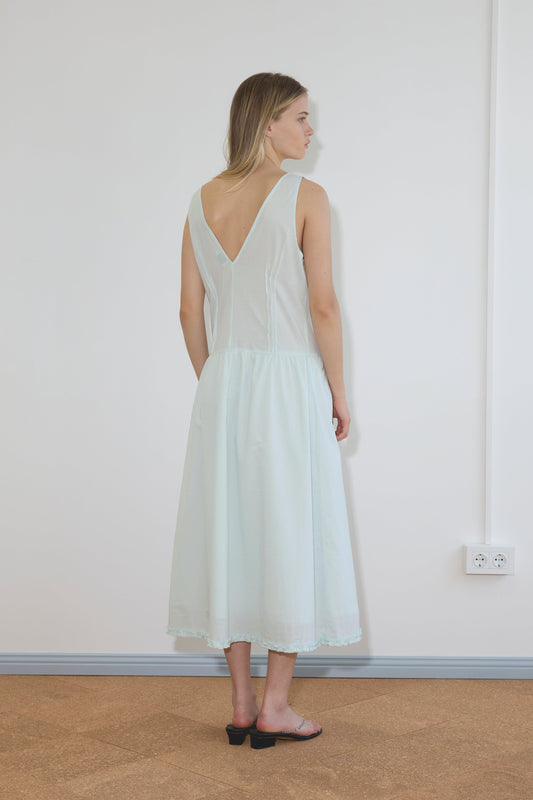 A woman with shoulder-length blonde hair and black sandals stands indoors, facing away, wearing the Pintuck Dress - Mint by Deiji Studios. The room has white walls, a carpeted floor, and a visible power outlet.