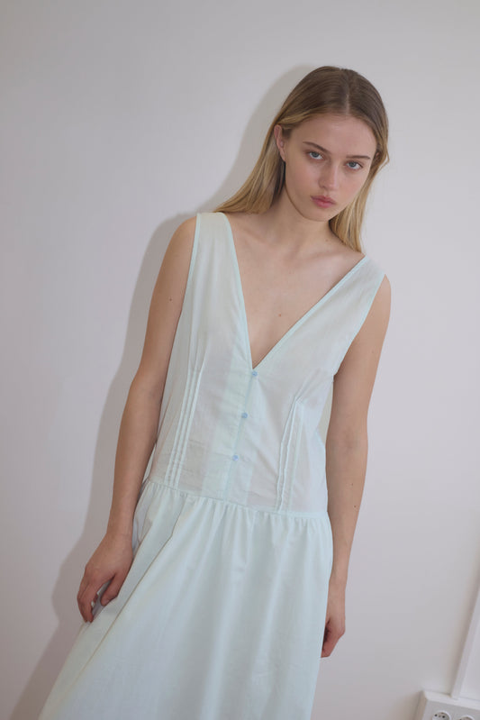 A woman with long blonde hair wears the Pintuck Dress - Mint by Deiji Studios, made from organic cotton, sleeveless and light blue-green, standing against a plain white wall and gazing slightly downward with a neutral expression.
