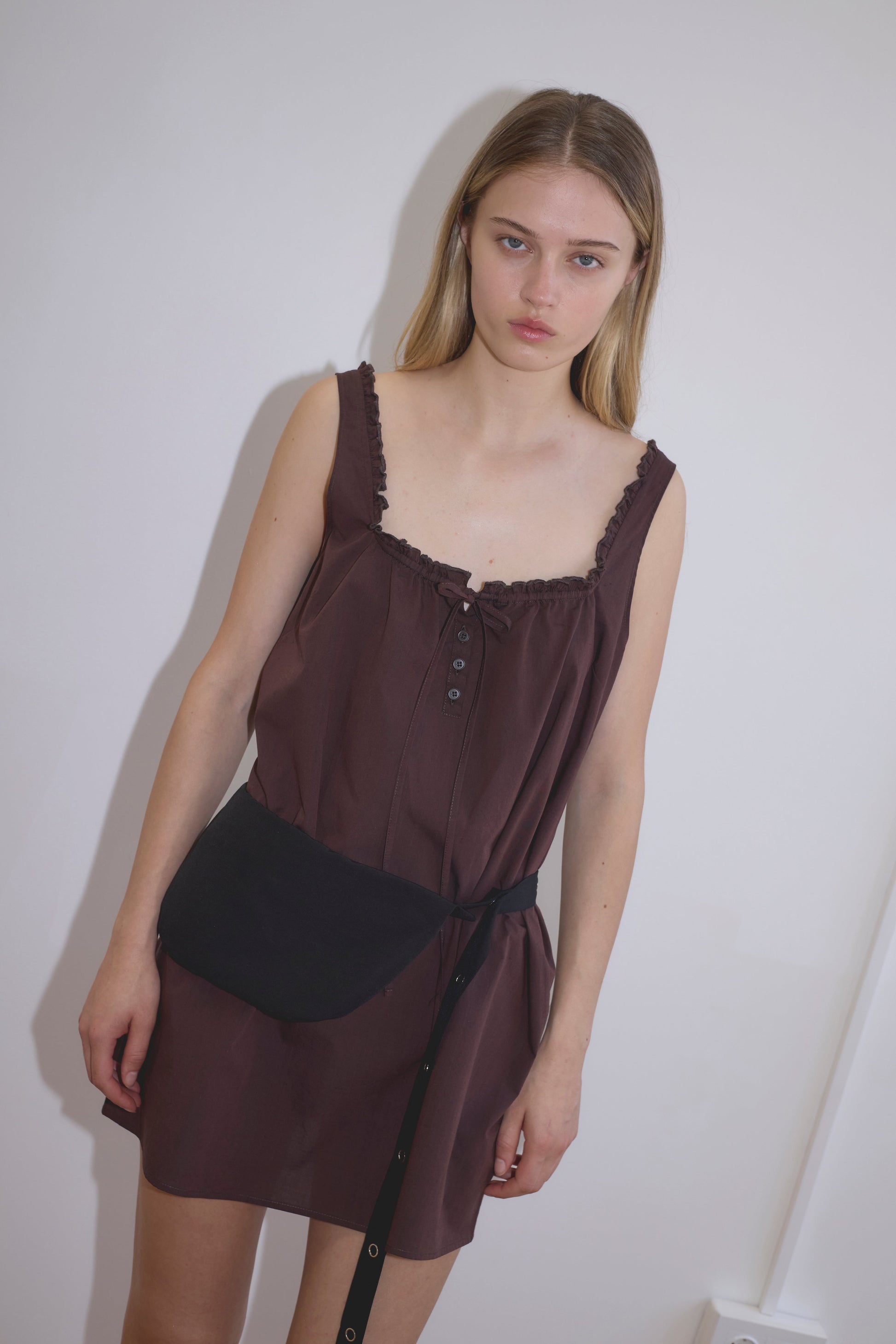 A young woman with long blonde hair wears the Button Frill Mini Dress, a sleeveless, dark brown dress with buttons and a ruffled neckline. She stands against a white wall with a black belt bag, looking at the camera neutrally.