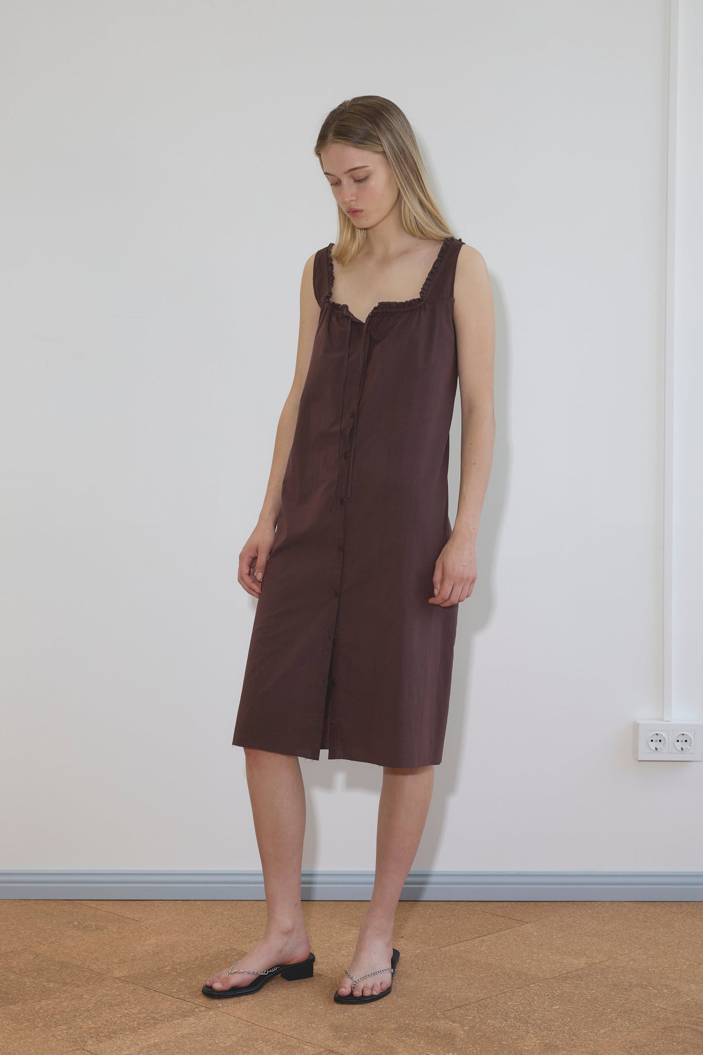 A woman stands indoors against a plain white wall, wearing the Button Sweep Dress—sleeveless, knee-length, button-down—and black sandals. She looks down with her arms relaxed at her sides.