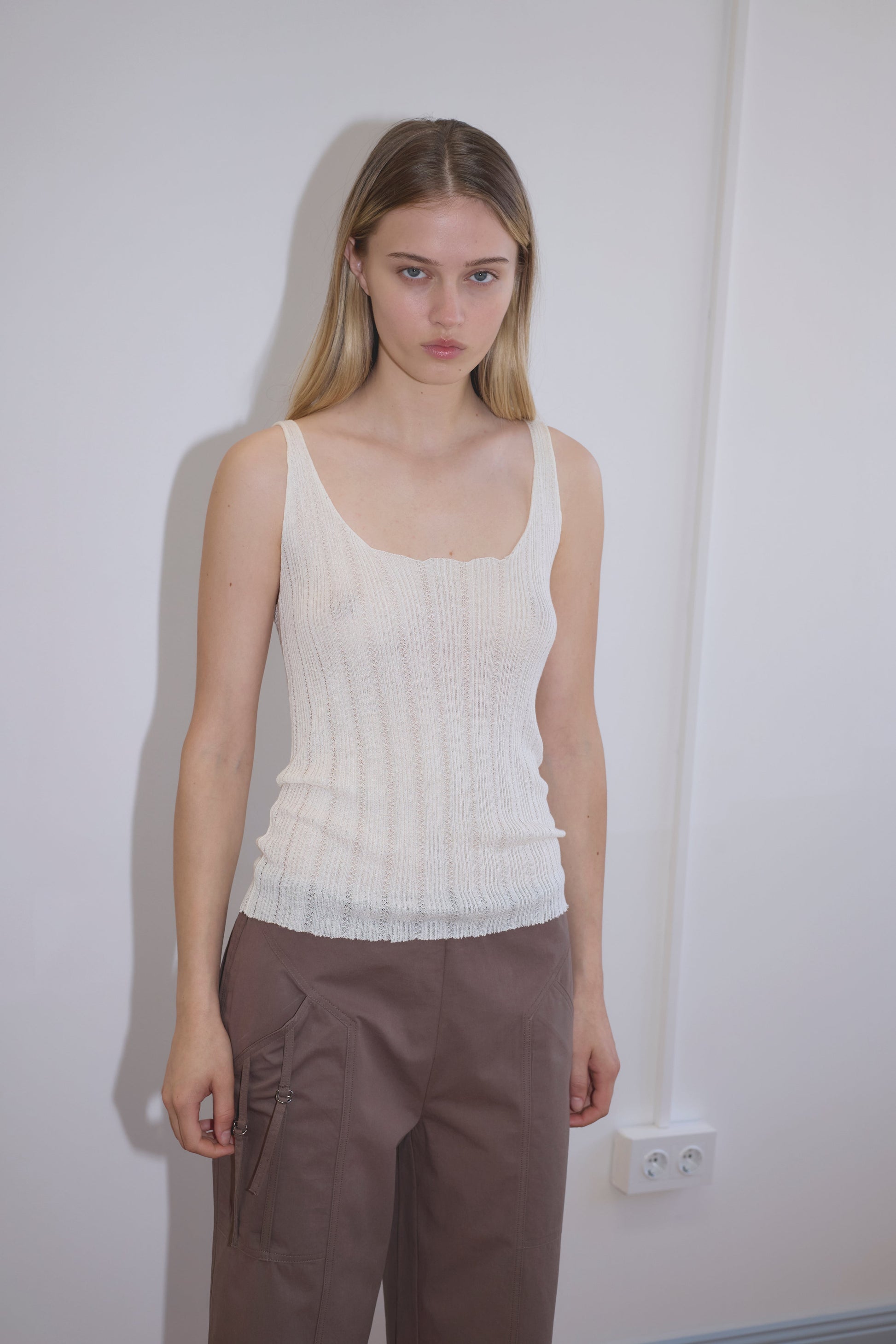 A young woman with straight blonde hair stands against a white wall, wearing the Open Work Tank - White by Deiji Studios and loose brown zipper pants, looking directly at the camera with a neutral expression.