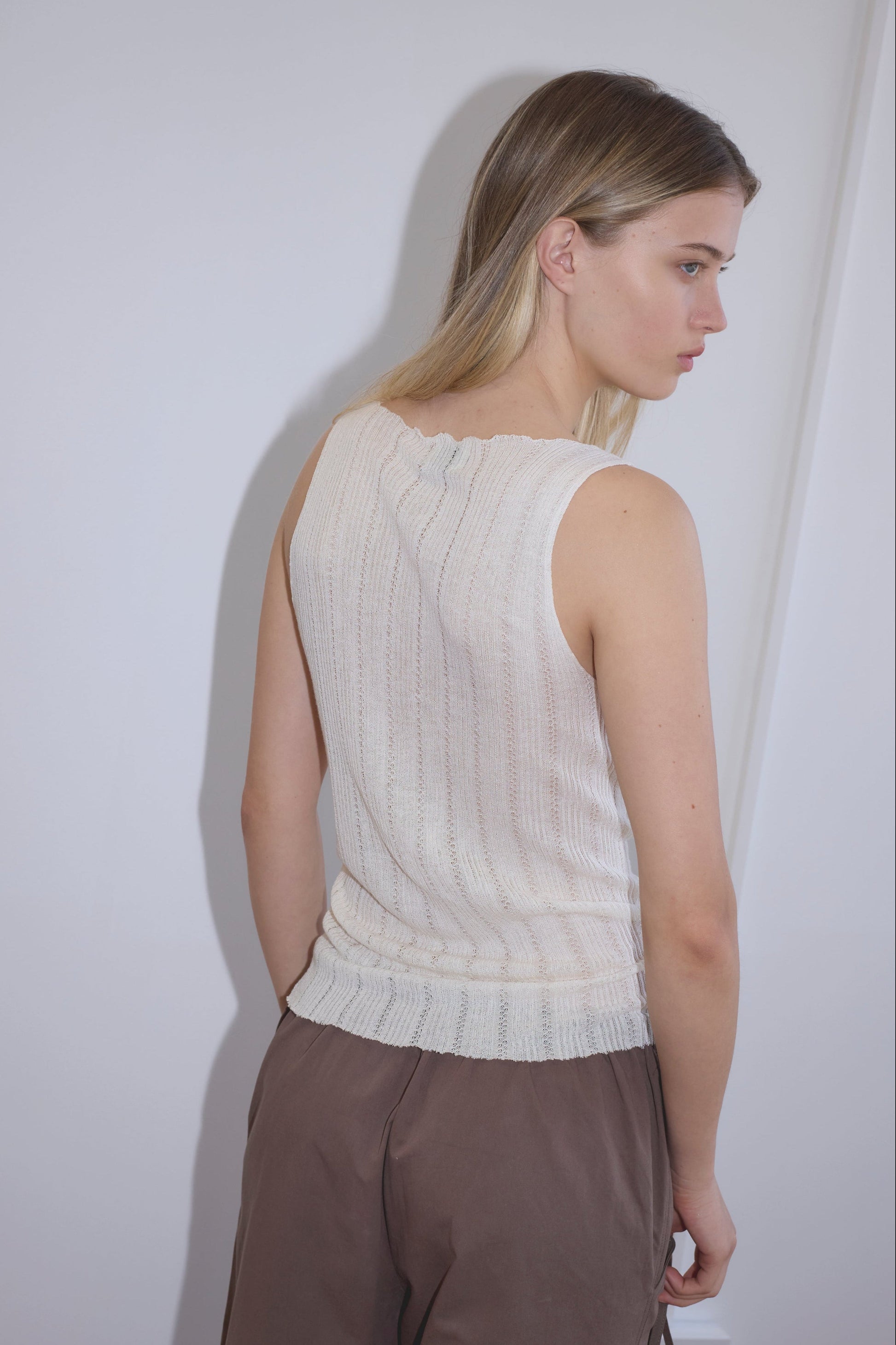 A woman with long blonde hair stands with her back turned and face in profile against a plain white wall, wearing the Open Work Tank - White by Deiji Studios and loose brown ecoviscose linen pants.