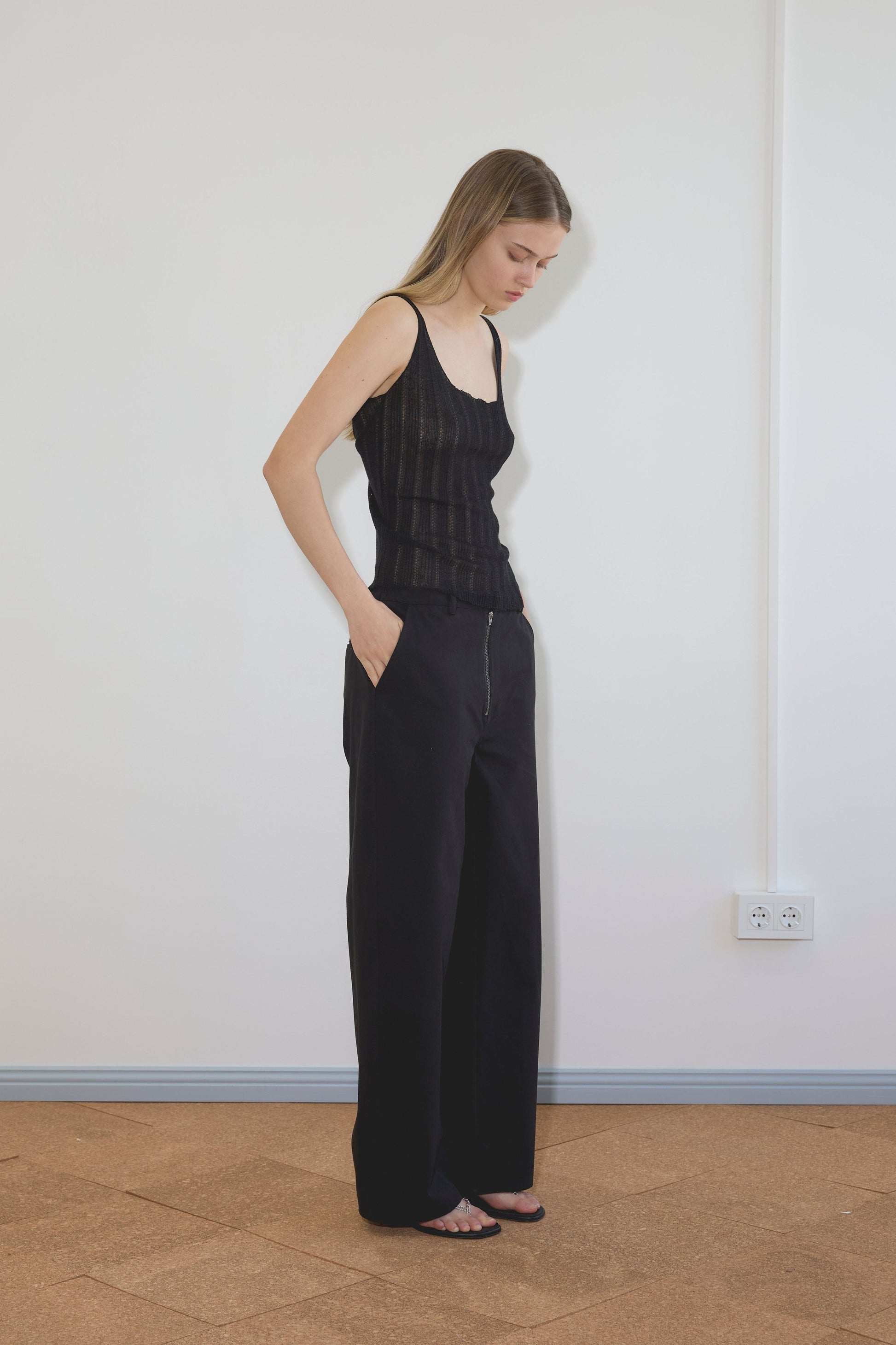 A woman with long hair stands indoors against a white wall, wearing the Deiji Studios Open Work Tank - Black, wide-leg black pants, and sandals. She has her hands in her pockets and looks down; an electrical outlet is visible on the wall.