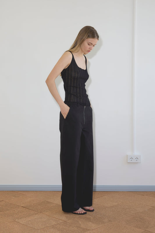 A woman with long hair stands indoors against a white wall, wearing the Deiji Studios Open Work Tank - Black, wide-leg black pants, and sandals. She has her hands in her pockets and looks down; an electrical outlet is visible on the wall.