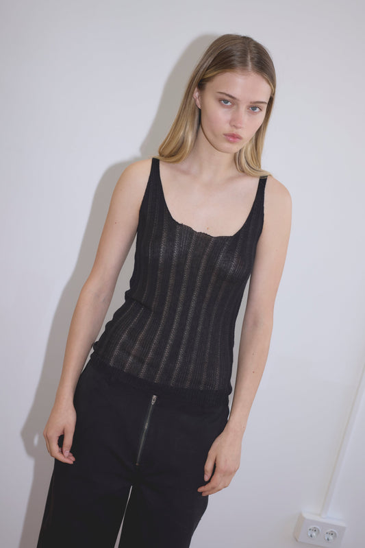 A young woman with straight blonde hair stands against a white wall, wearing the Open Work Tank - Black by Deiji Studios and black pants. She gazes slightly to the side with a neutral expression.