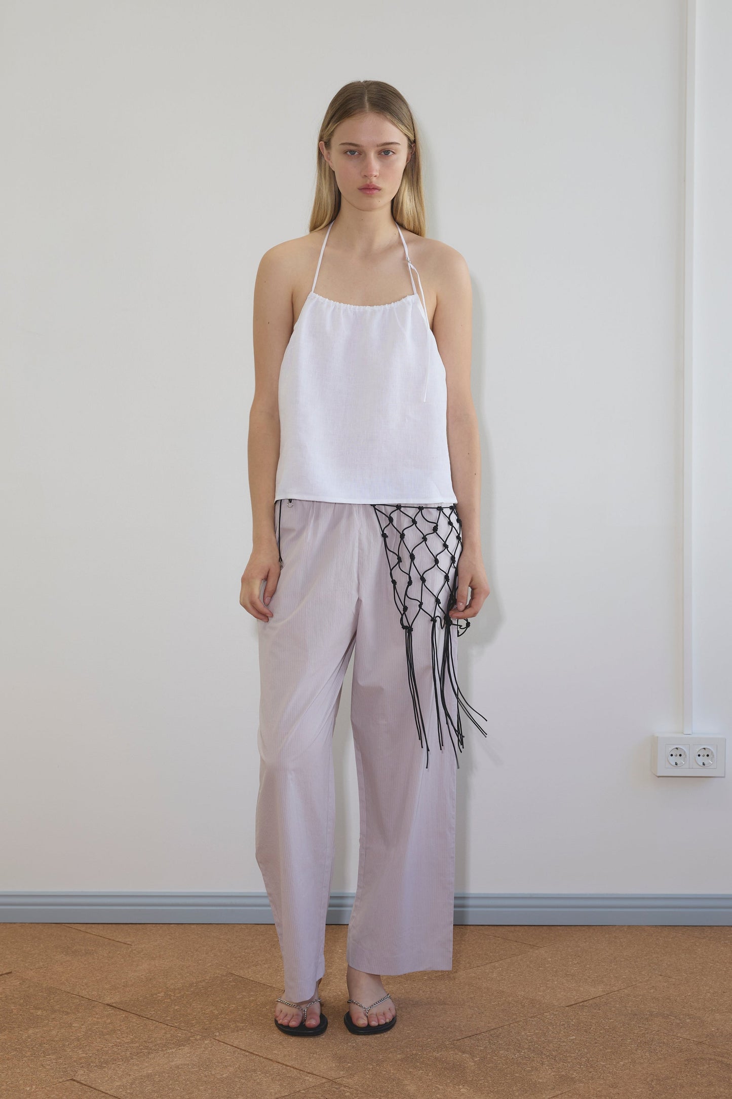 Model wearing a white linen halter top and Ease Trouser - Lilac Stripe by Deiji Studios, paired with a black macramé net bag and silver chain flip-flops, standing against a white wall.