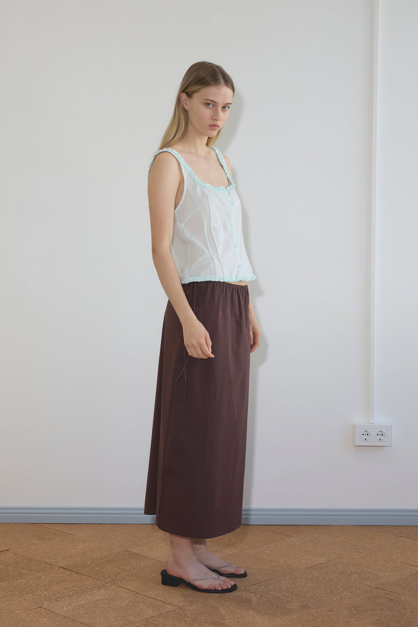 A woman stands indoors against a white wall, wearing the Deiji Studios Button Frill Top in Mint with a long brown skirt and black sandals, looking at the camera with a neutral expression.