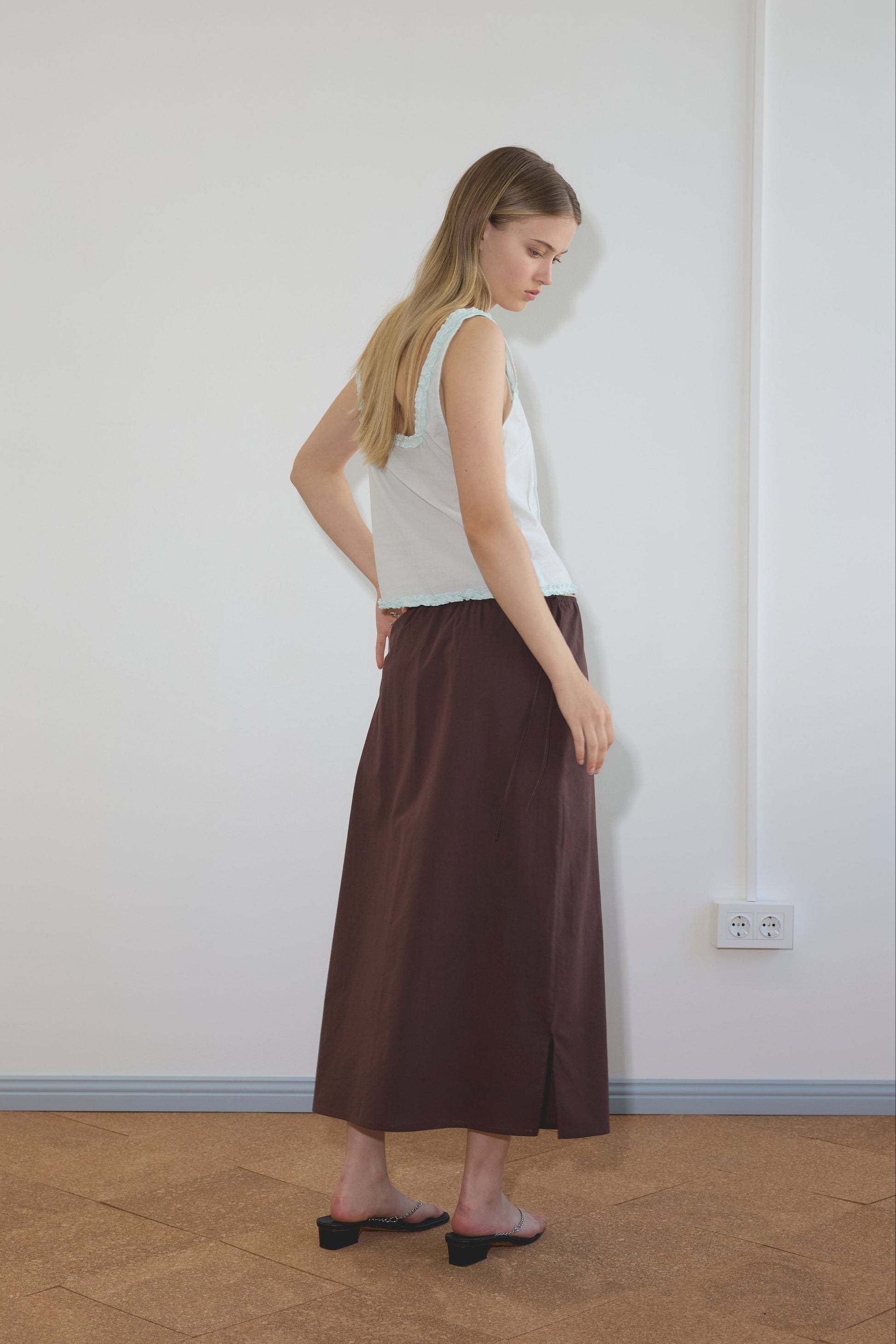 A woman with long blonde hair stands sideways in a minimalist room, wearing Deiji Studios’ Button Frill Top in Mint, a long brown skirt, and black open-toe shoes on a wooden floor with white walls.