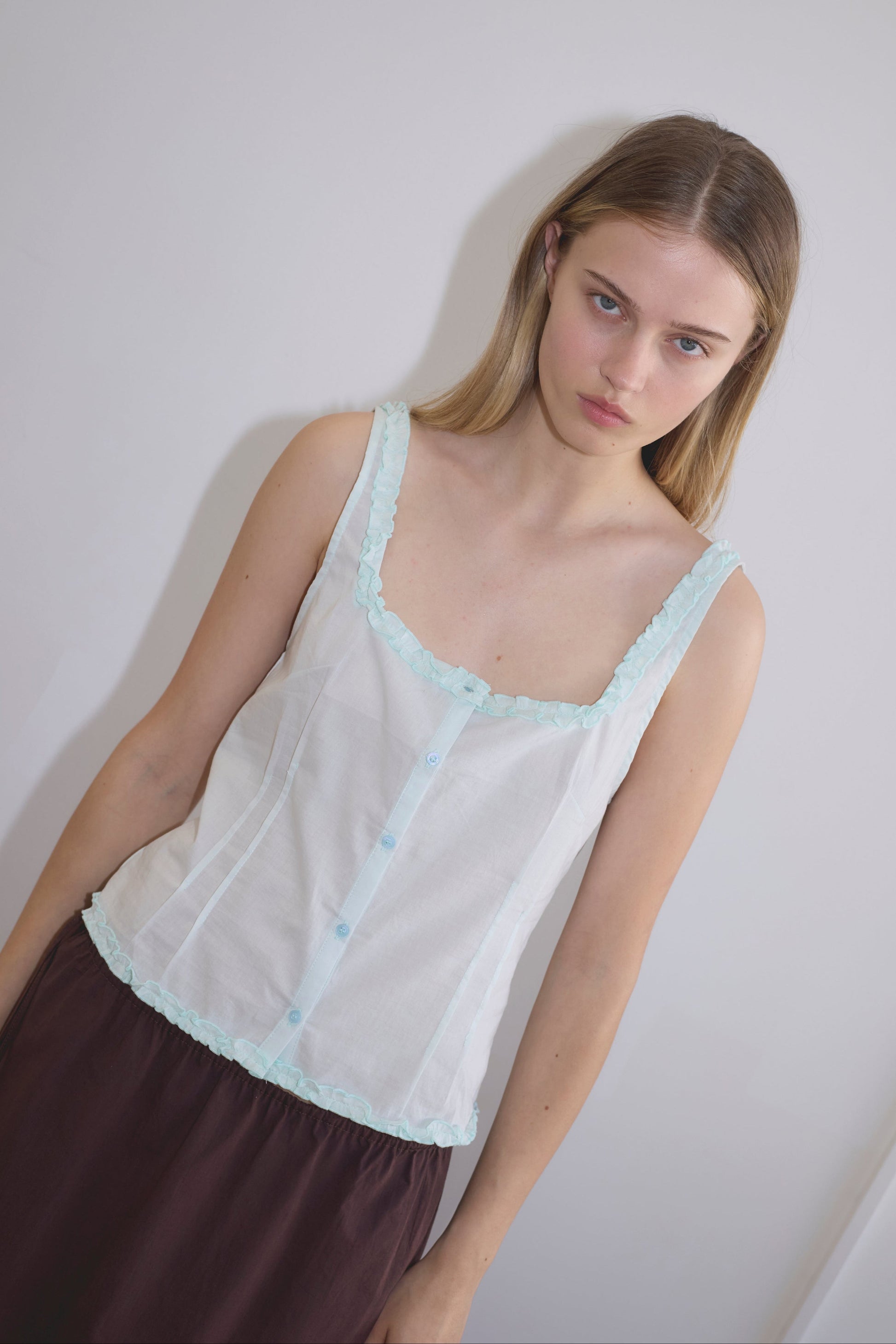A young woman with long blonde hair wears the Button Frill Top - Mint by Deiji Studios, featuring a ruffled square neckline and light blue trim, paired with dark brown pants, standing against a plain, light-colored wall.