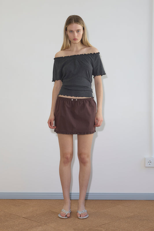 A woman stands indoors against a white wall, wearing an off-the-shoulder dark gray top, the Flounce Mini - Umber skirt by Deiji Studios with an elastic waistband, and sandals. She has straight long blonde hair and a neutral expression.