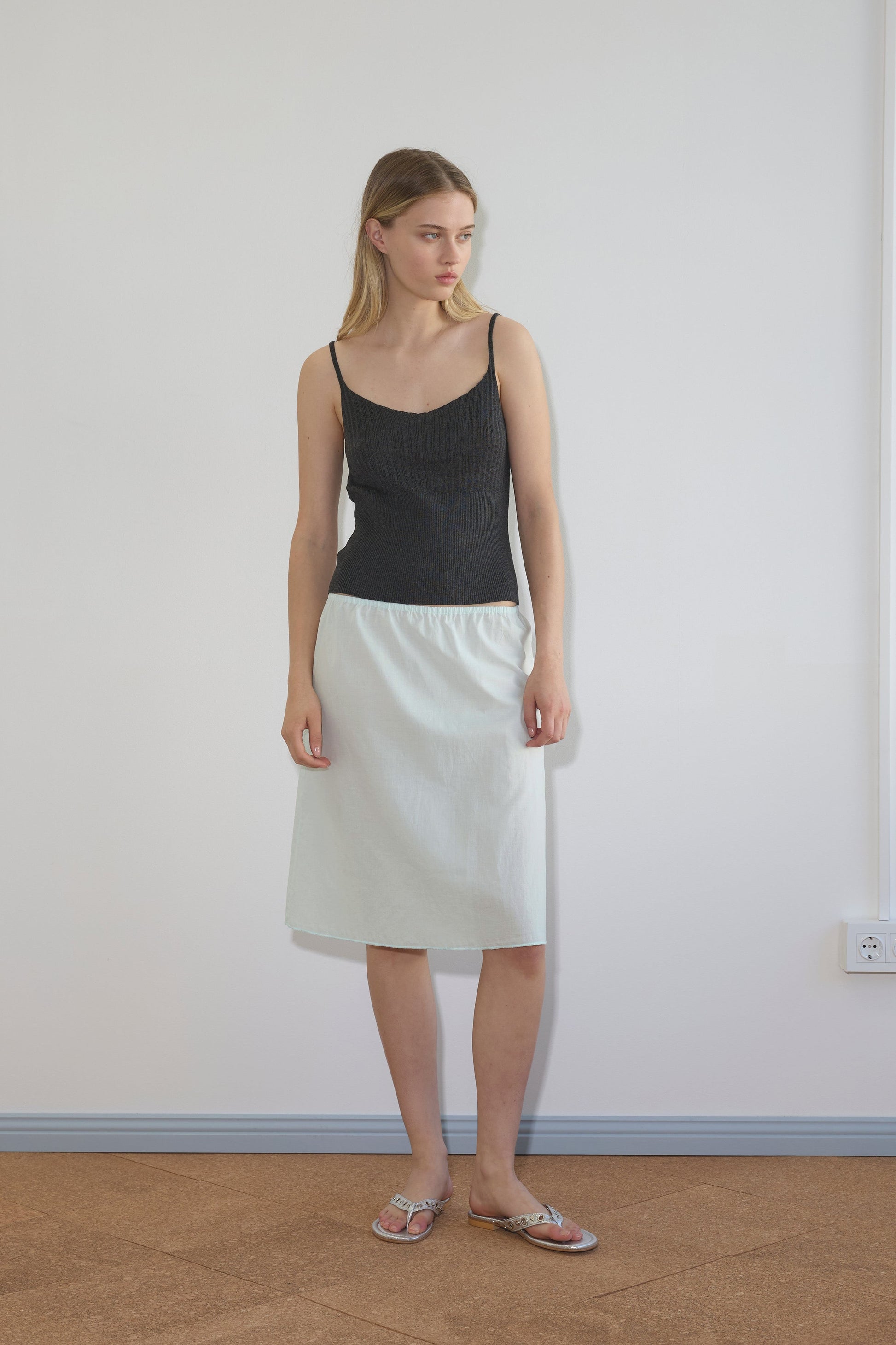 A woman stands indoors against a plain white wall, wearing a black sleeveless top and the Simple Skirt - Mint from Deiji Studios. She looks slightly to the side with a neutral expression, her outfit completed by simple sandals.