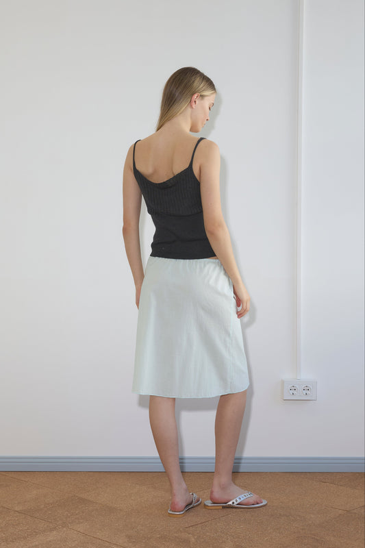 A woman stands indoors against a white wall, wearing a black sleeveless top and the Simple Skirt - Mint. She has light hair, gazes slightly left, and an electrical outlet appears on the wall.
