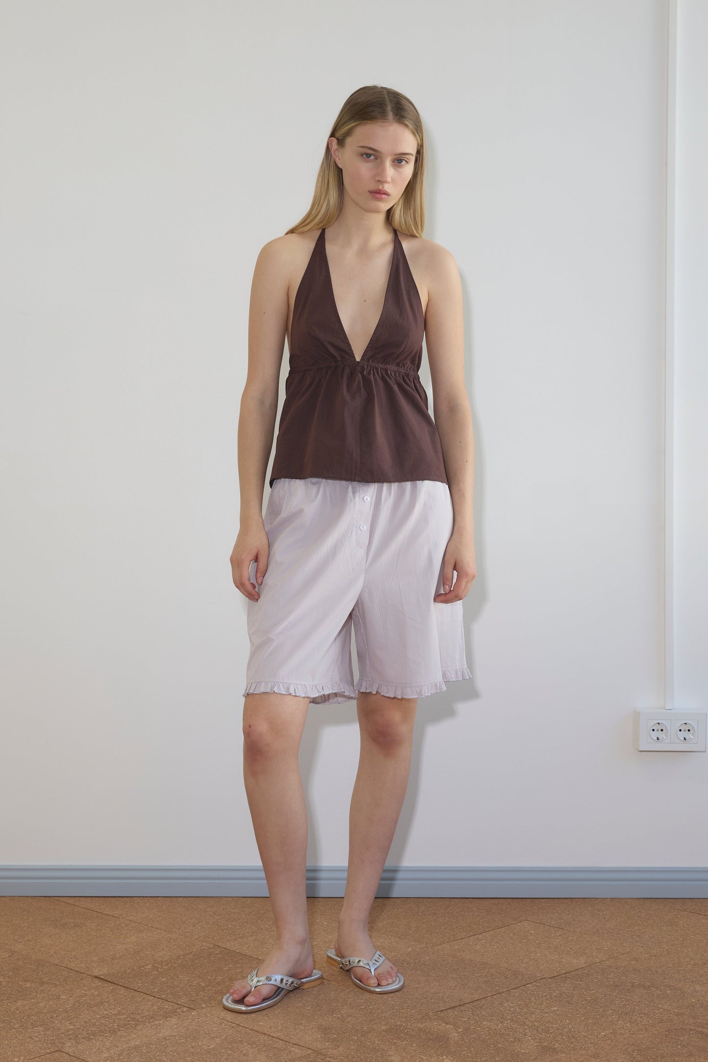 A young woman with straight light brown hair and a neutral expression stands indoors against a plain white wall, wearing loose Flounce Shorts, a sleeveless brown halter top, and sandals.