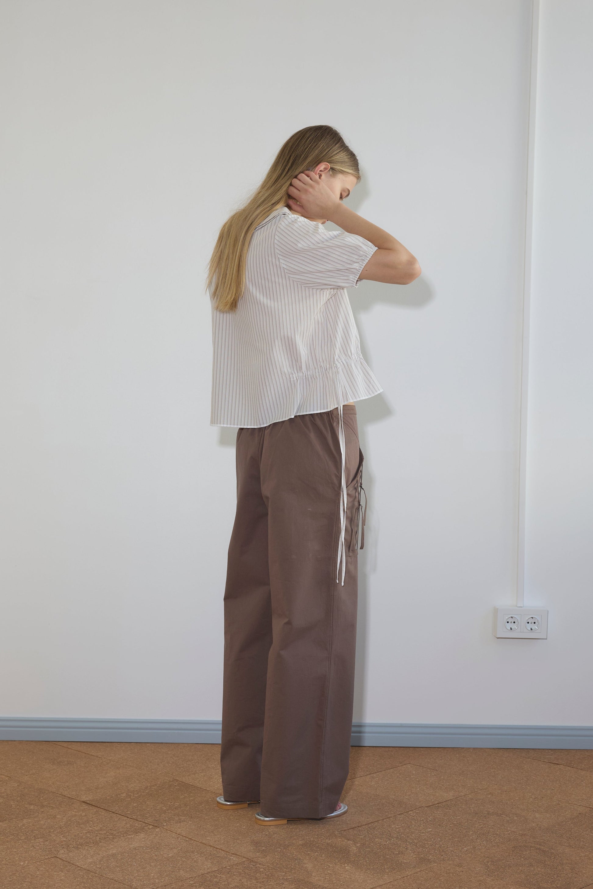 A woman with long blonde hair faces a white wall, wearing the Double Collar Shirt—a loose, short-sleeved, white striped blouse—paired with wide-legged brown pants. She holds her neck with one hand and slightly bows her head.