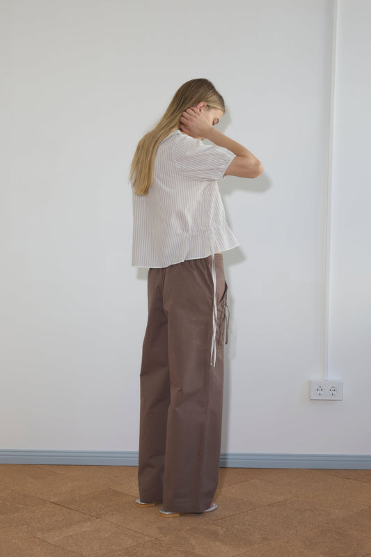 A woman with long blonde hair faces a white wall, wearing the Double Collar Shirt—a loose, short-sleeved, white striped blouse—paired with wide-legged brown pants. She holds her neck with one hand and slightly bows her head.