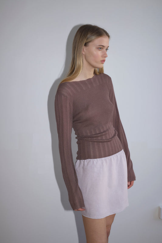 A woman with long blond hair stands against a light gray wall, wearing a fitted brown top and the Babylock Skirt, a short white skirt, looking to her left with a neutral expression.