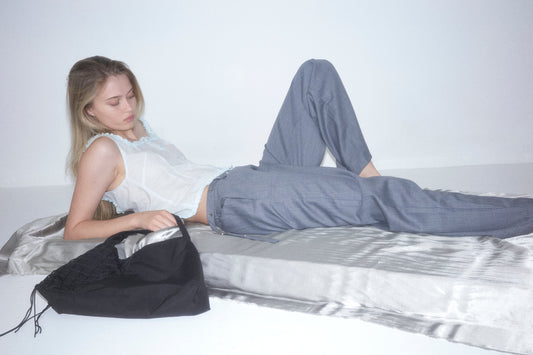 A blonde woman reclines on silky silver fabric in a sheer white camisole and gray pants. Next to her is the Deiji Studios Net Contrast Bag - Black, made from recycled polyester, set against a plain white background.