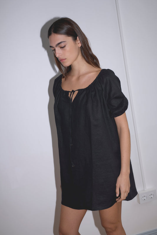 A woman with long brown hair wears the Smock Dress - Black by Deiji Studios—loose-fitting with puffed sleeves—standing against a white wall and looking down with a neutral expression.