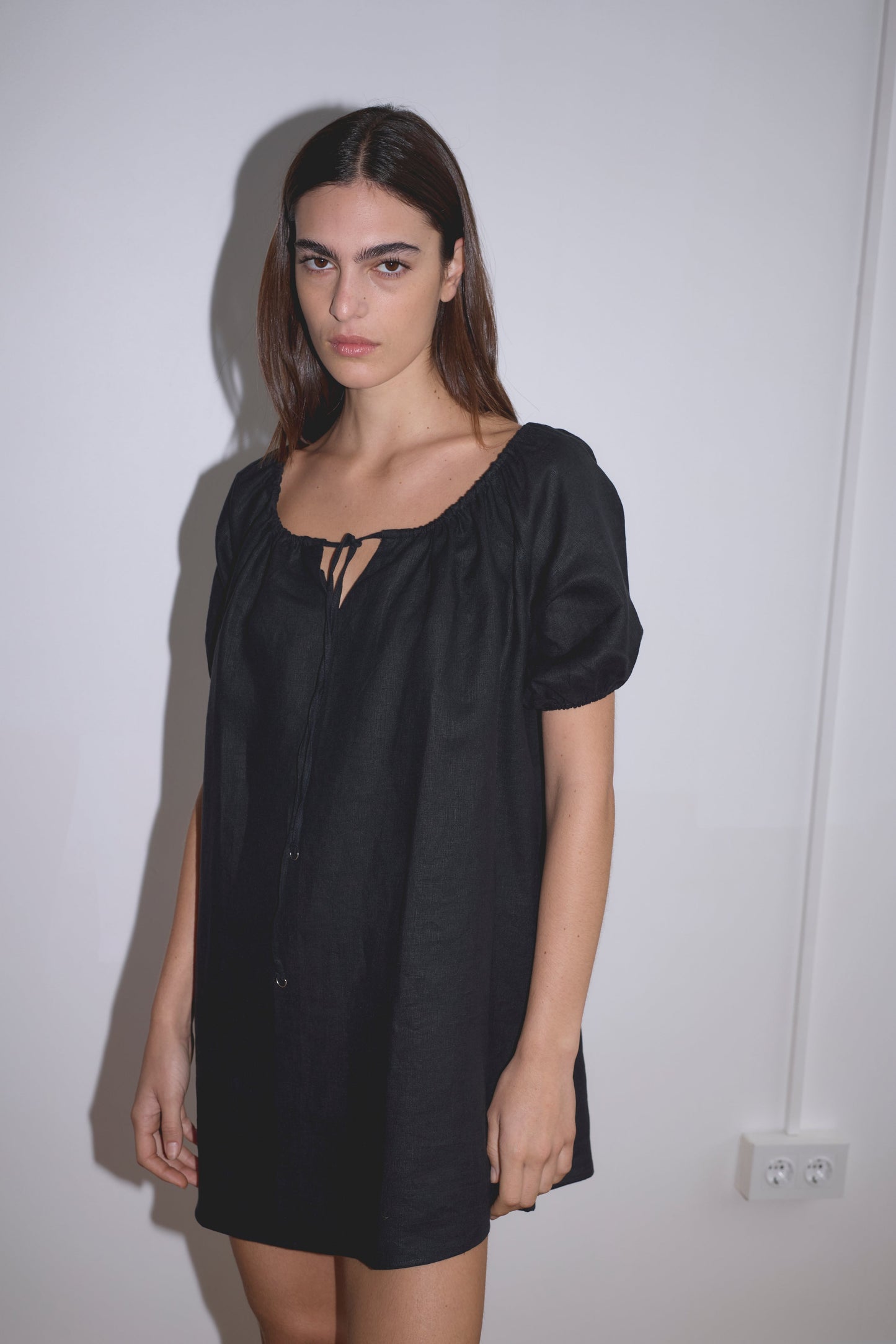 A person with long brown hair stands against a white wall, wearing the Smock Dress - Black, a loose-fitting French linen dress with short sleeves and a tie neckline, evoking Deiji Studios’ iconic style.