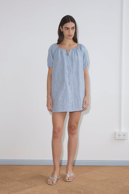 A woman stands indoors against a white wall, wearing the Smock Dress - Blue Stripe, a short dress with short sleeves and a front tie detail. She has long brown hair, a neutral expression, and is wearing sandals.