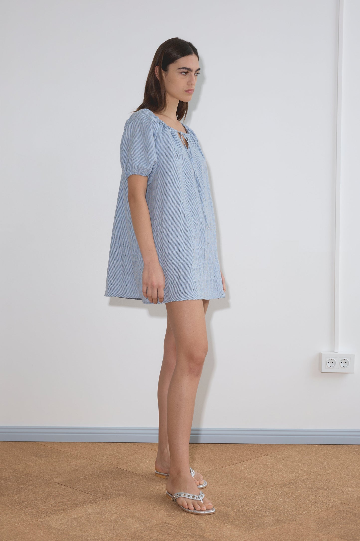 A woman with straight brown hair stands indoors against a white wall, wearing the Smock Dress - Blue Stripe and clear sandals. She looks ahead with a neutral expression near an electrical outlet.