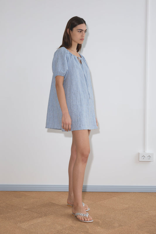 A woman with straight brown hair stands indoors against a white wall, wearing the Smock Dress - Blue Stripe and clear sandals. She looks ahead with a neutral expression near an electrical outlet.